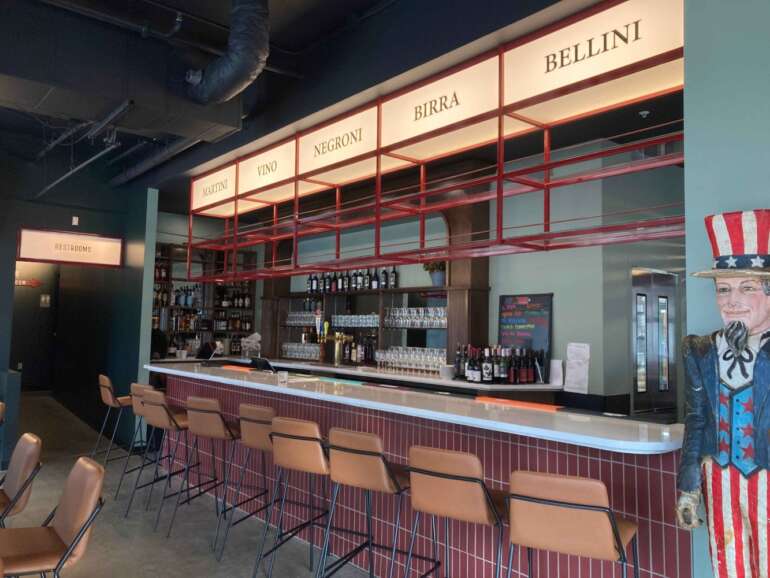The Spaghetti Warehouse Has A New Location In Downtown Columbus 6 Modern bar interior featuring illuminated signage for martini, vino, negroni, birra, and bellini drinks above counter with tan bar stools.