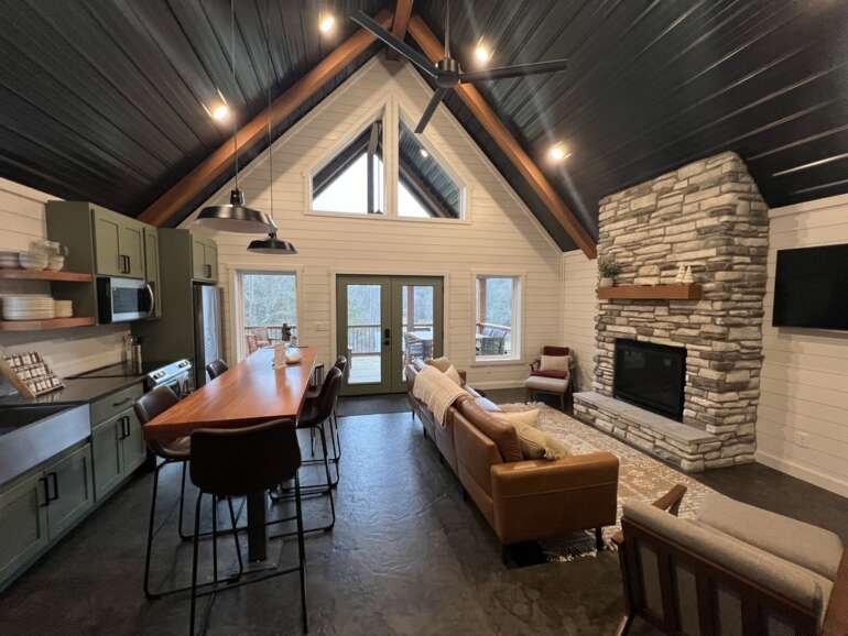 Modern open-concept cabin interior with kitchen, fireplace, and vaulted ceiling with exposed beams.