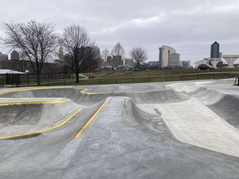 Concrete skate park with yellow line markings and city skyline visible in the background under cloudy sky.