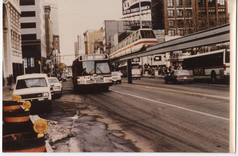 Downtown street scene with elevated transit train, buses, and construction barriers along urban roadway.