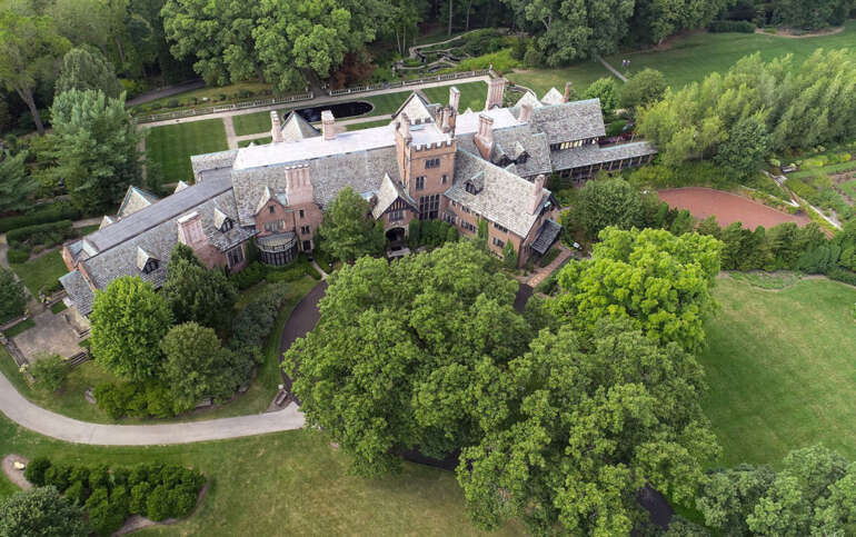 Aerial view of a brick manor house with slate roofs surrounded by manicured grounds and dense green trees.