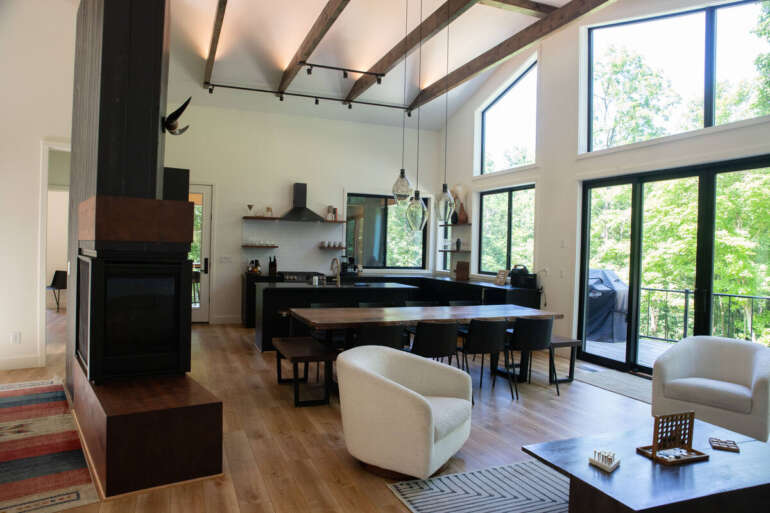From Cozy Comfort to Gorgeous Grounds: The Acres Cabin Has It All 10 Modern open-concept home interior featuring black kitchen island, cream armchairs, exposed beams, and expansive windows overlooking wooded landscape.