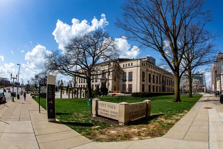 Wide-angle view of Columbus City Hall exterior with bare trees, green lawn, and stone signage on sunny day.