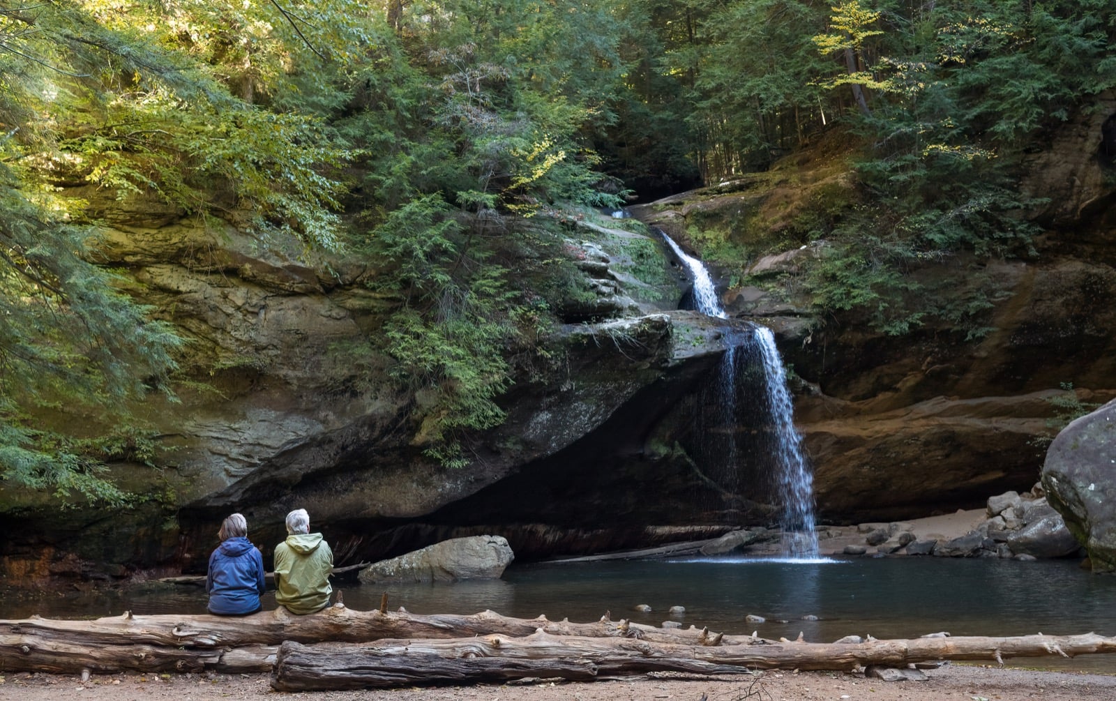 Waterfall at Lower Falls at Old Man's Cave in Hocking Hills State Park, with two hikers sitting on a log by the pool