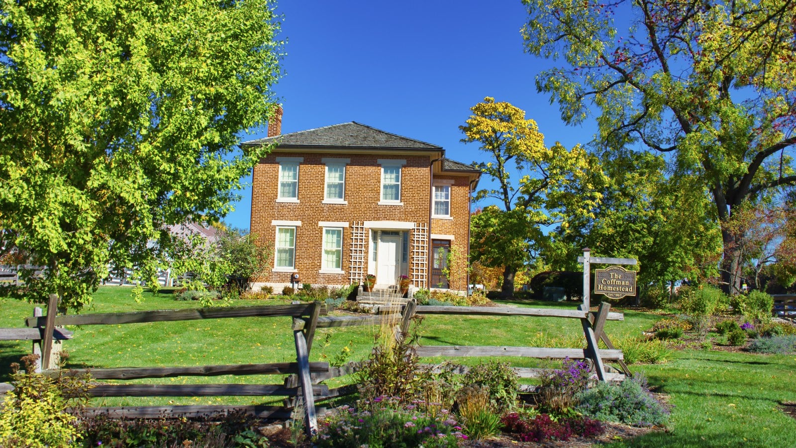 Two-story brick Coffman farmhouse behind a split-rail fence and flower beds, with a hanging sign reading The Coffman Homestead