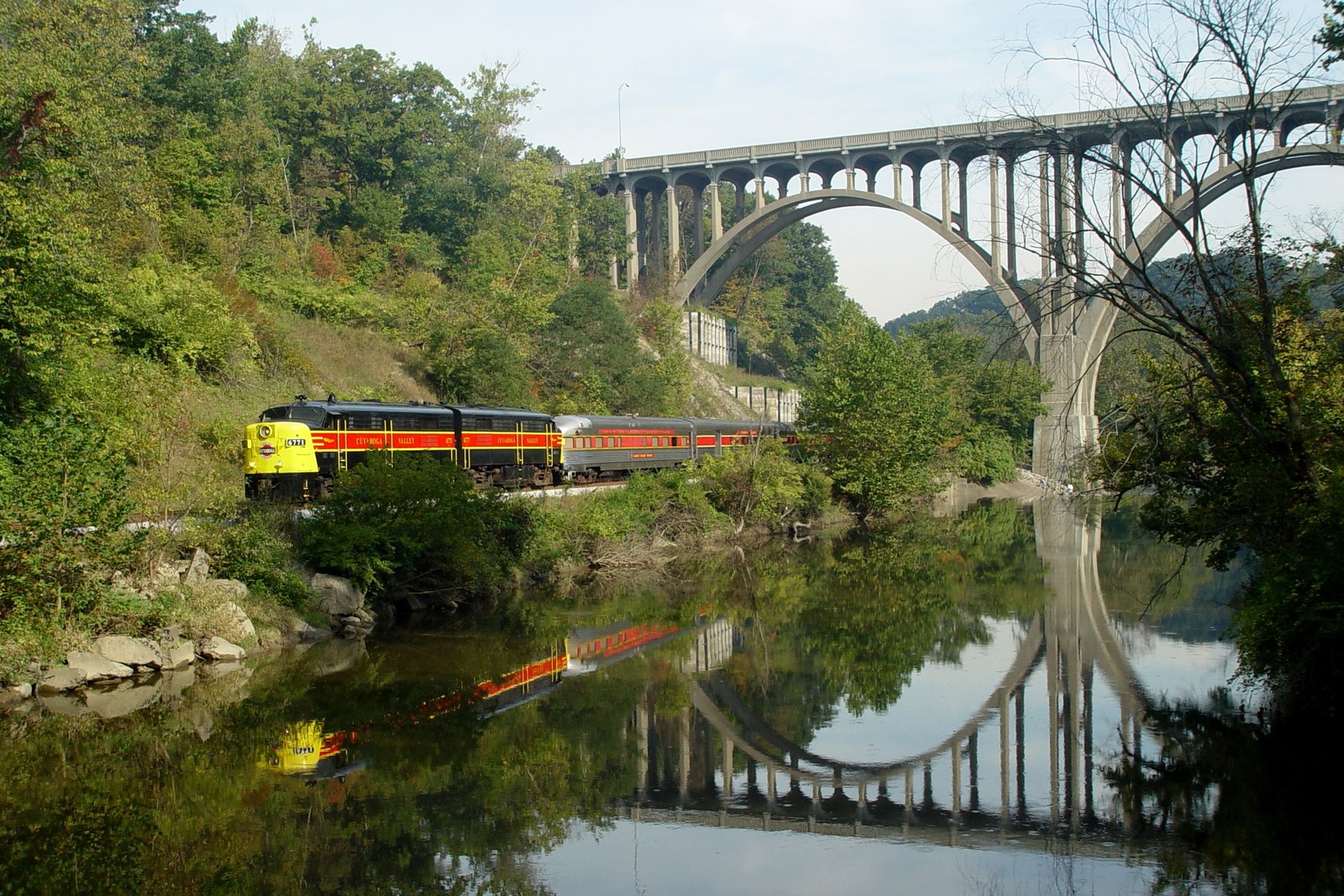 Cuyahoga Valley National Park Belongs On Your Fall Bucket List 2 A yellow and red Cuyahoga Valley Scenic Railroad train runs along the Cuyahoga River beneath a concrete arch bridge, both reflected in the still water.