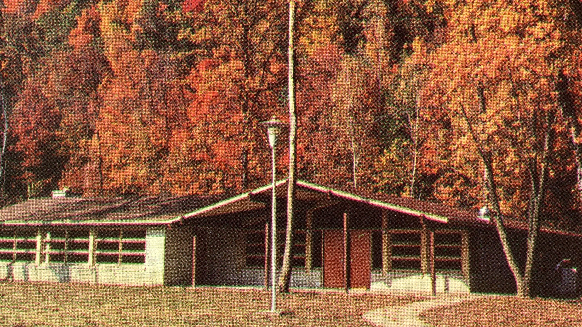 The 8 Best Places In Ohio To See Fall Foliage 3 Red and orange fall trees on a hillside behind a low park building at Mohican State Park on a vintage postcard