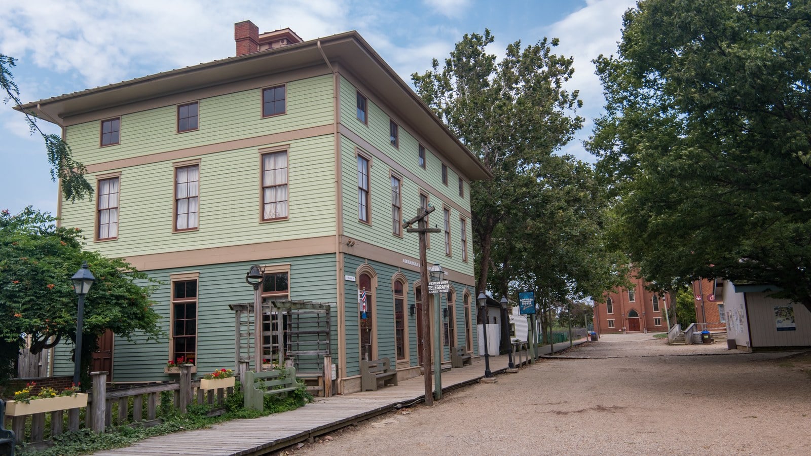 Green clapboard American House Hotel on Ohio Village's gravel main street, with a wooden boardwalk and a brick church at the far end